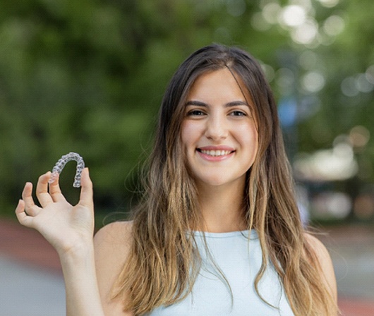 A teenage girl wearing a sleeveless, white shirt holds an Invisalign aligner in her right hand and smiles